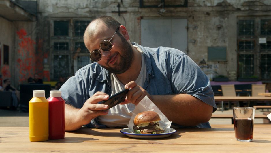 Smiling man taking a photo of a burger with a smartphone at an outdoor café.