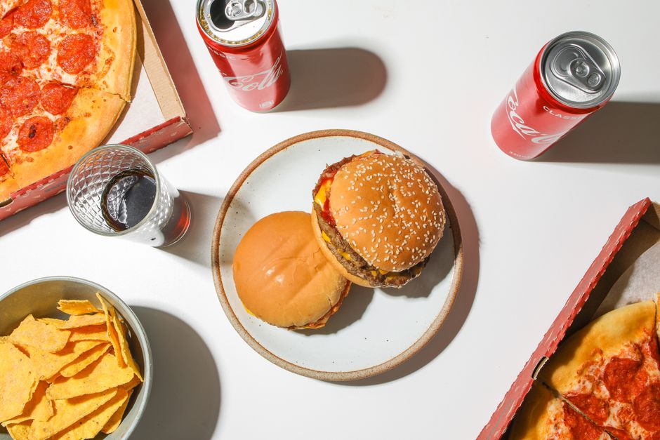 An appetizing fast food spread featuring pizza, burgers, and cola cans on a white tabletop.