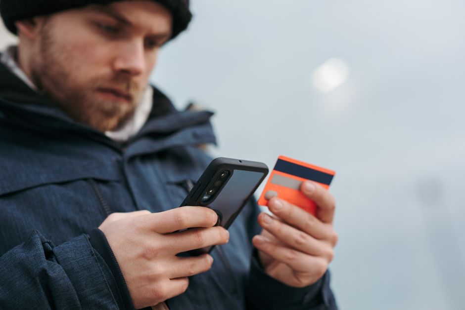 Crop male in outwear entering details of credit card on mobile phone while making online payment for purchase in daytime